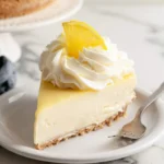 A creamy slice of No-Bake Lemon Cheesecake topped with whipped cream and a lemon slice on a white plate.
