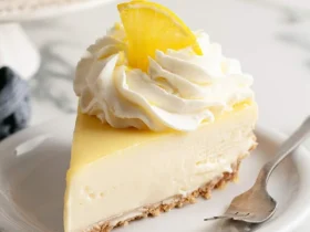 No-Bake Lemon Cheesecake 50 A creamy slice of No-Bake Lemon Cheesecake topped with whipped cream and a lemon slice on a white plate.